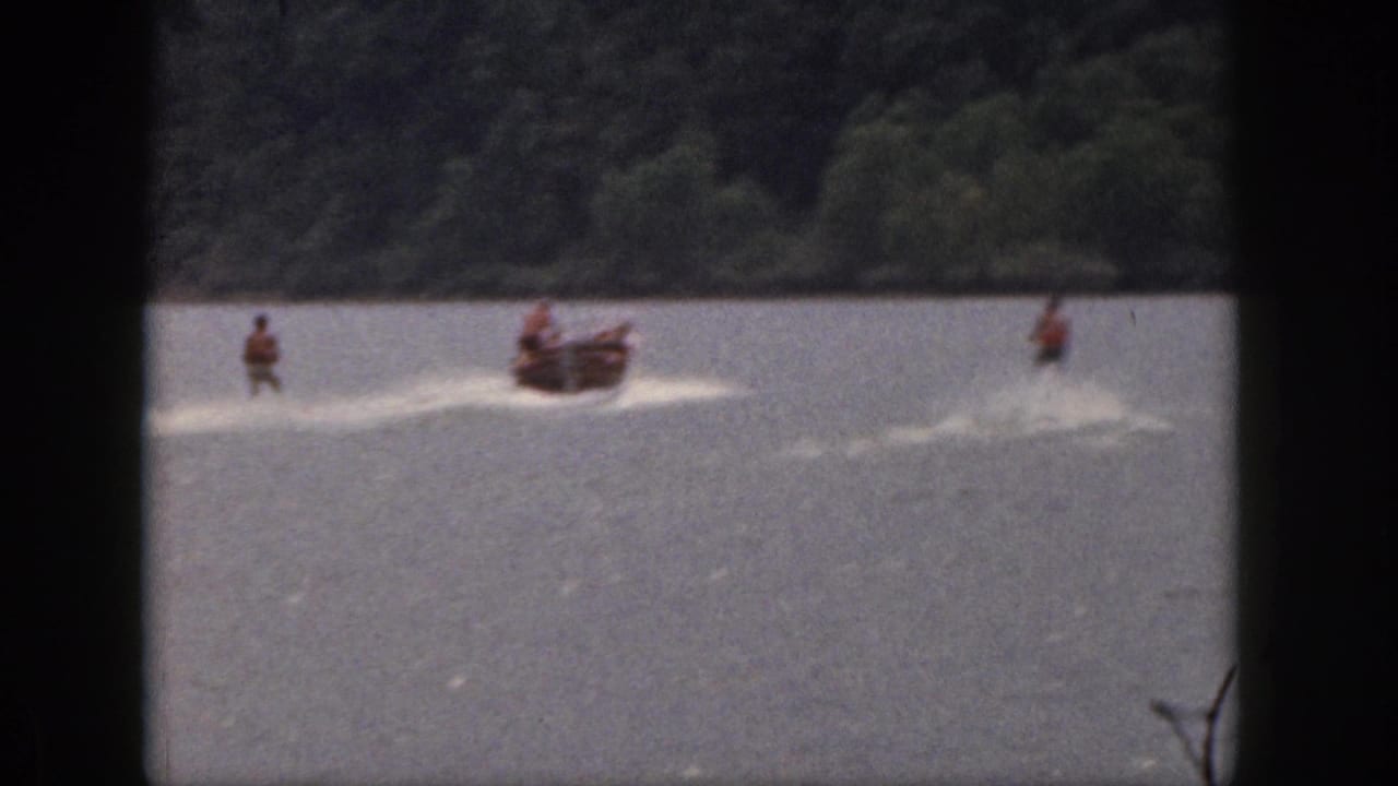 1961 Michigan home-movie frame showing a motorboat towing multiple water skiers across open lake water near trees.