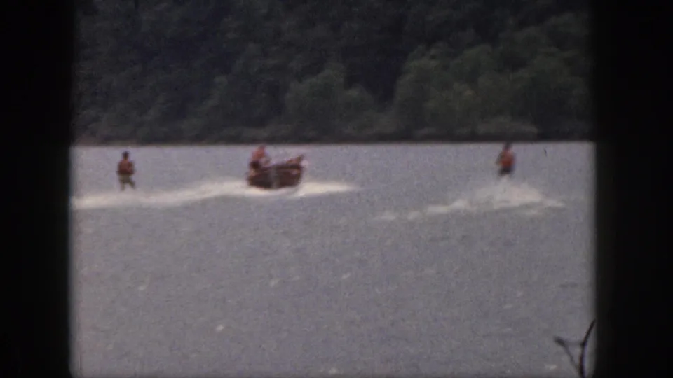 1961 Michigan home-movie frame showing a motorboat towing multiple water skiers across open lake water near trees.