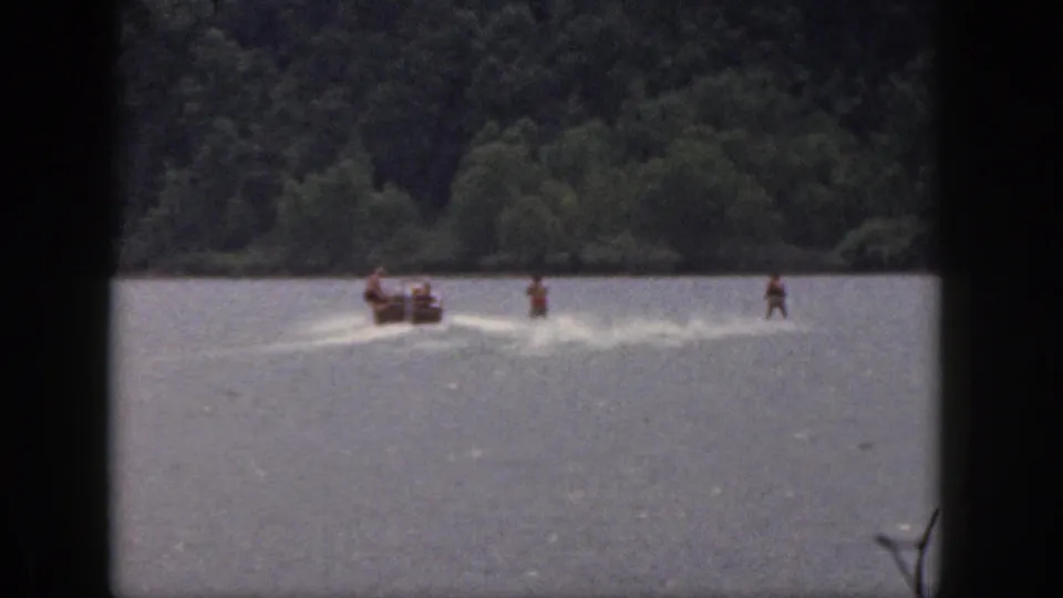 1961 Michigan home-movie view of several water skiers spread behind a motorboat on a wooded lake.