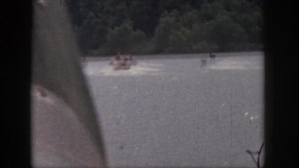 1961 Michigan 8mm home-movie view of a motorboat pulling waterskiers across a wooded lake, partly obscured by the film frame.