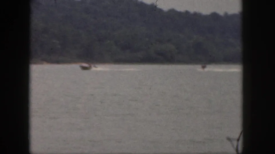 Soft 1961 Michigan home-movie frame showing a tow boat and distant waterskiers moving across a lake below a dense treeline.
