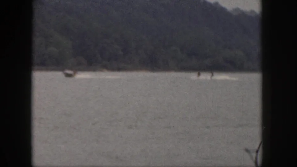 Grainy 8mm frame of distant waterskiing on a Michigan lake, with a tow boat, two small skier figures, and a wooded backdrop.