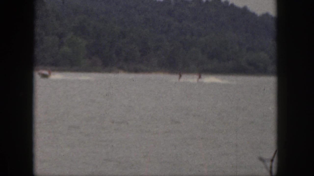 1961 Michigan home-movie frame showing two distant water skiers pulled behind a small motorboat on a lake with wooded shoreline beyond.