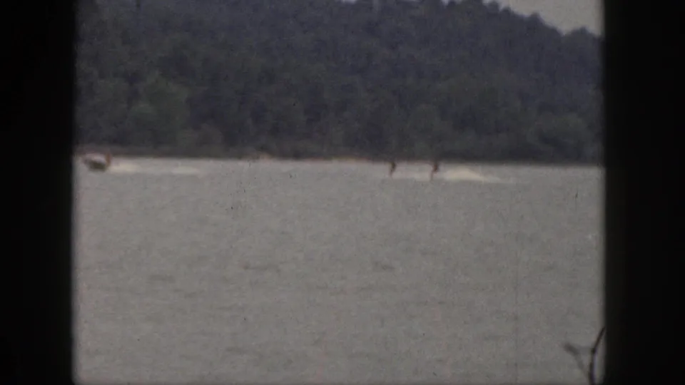 1961 Michigan home-movie frame showing two distant water skiers pulled behind a small motorboat on a lake with wooded shoreline beyond.