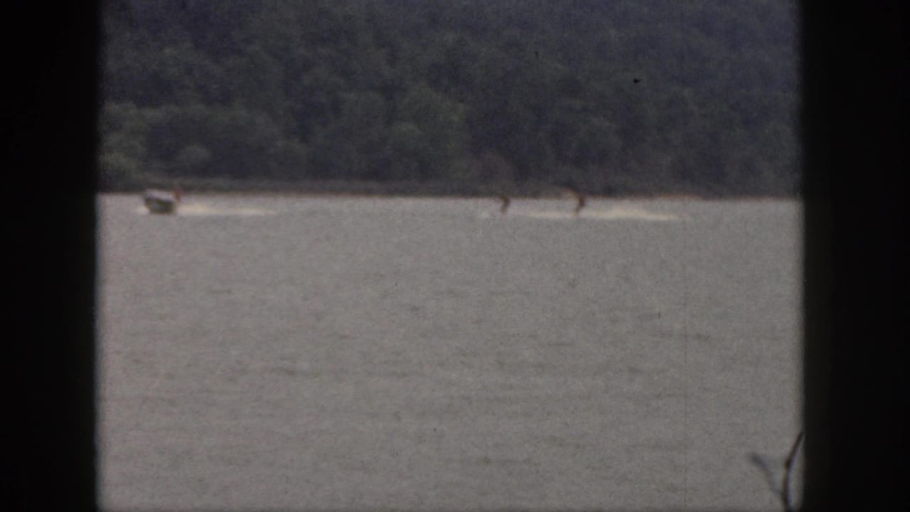 A motorboat pulls two distant skiers across open water with trees along the far shore.