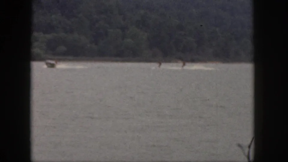 1961 Michigan 8mm frame with a small boat towing two water skiers across a calm lake below a tree-lined hillside.