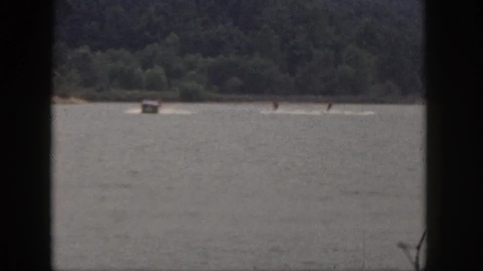1961 Michigan home-movie frame showing a small boat and two distant skiers leaving white wakes near a wooded lakeshore.