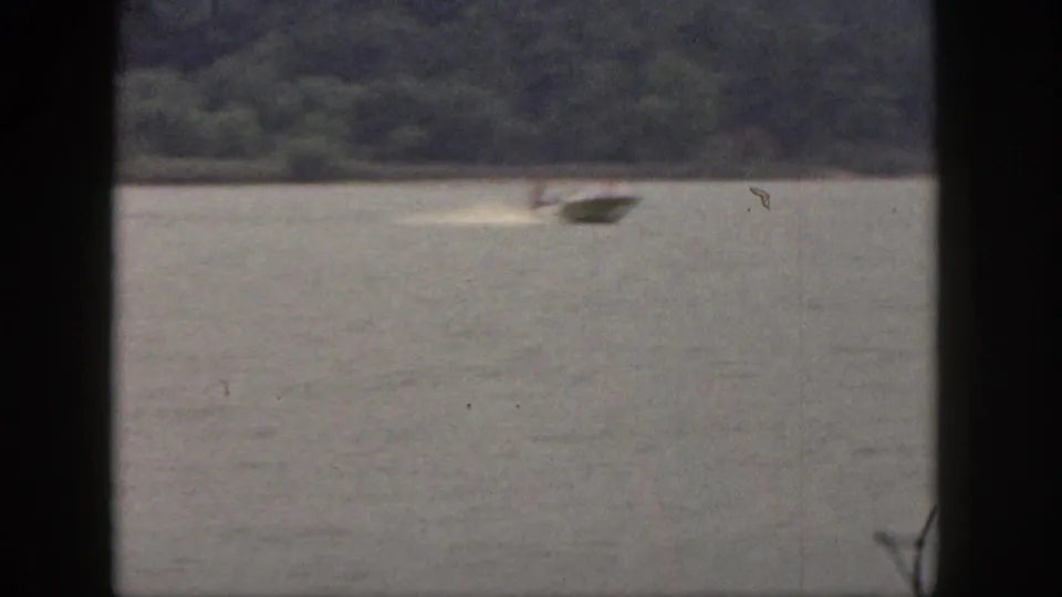 1961 Michigan 8mm frame with a motorboat crossing open water and distant water-ski activity near a wooded shoreline.