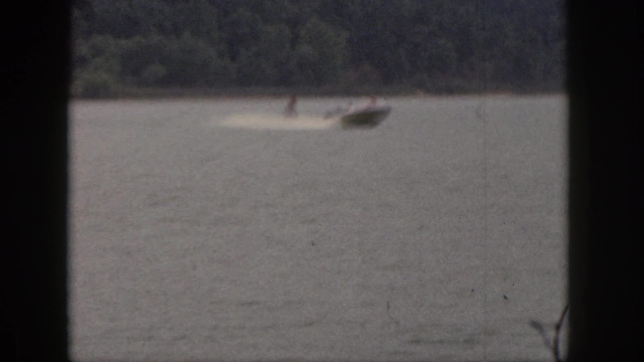 1961 Michigan home-movie frame showing a distant man water skiing behind a small speedboat on a tree-lined lake.