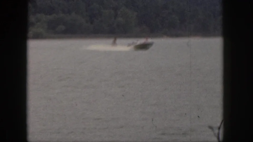 1961 Michigan home-movie frame showing a distant man water skiing behind a small speedboat on a tree-lined lake.