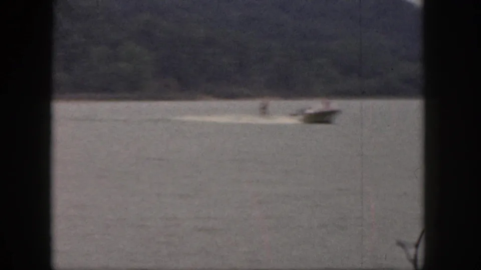 A small boat moves across a Michigan lake in 1961, pulling a distant water skier with a wooded shoreline behind them.