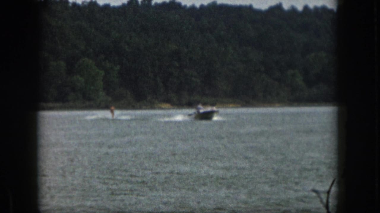 1961 Michigan 8mm frame showing a distant motorboat towing a water skier across a calm lake with trees in the background.