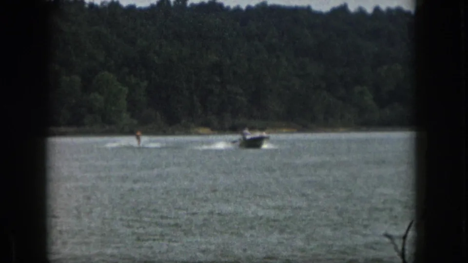 1961 Michigan 8mm frame showing a distant motorboat towing a water skier across a calm lake with trees in the background.