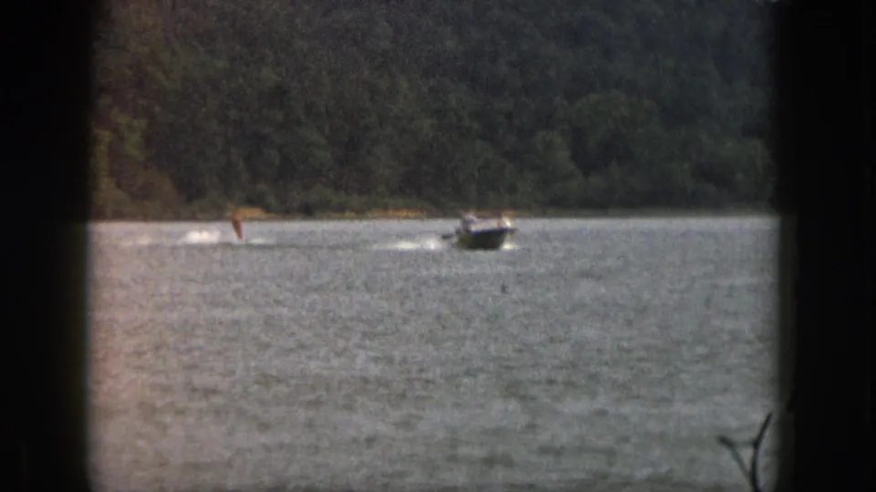 1961 Michigan 8mm frame with a small motorboat towing a distant skier across open water below a wooded shoreline.