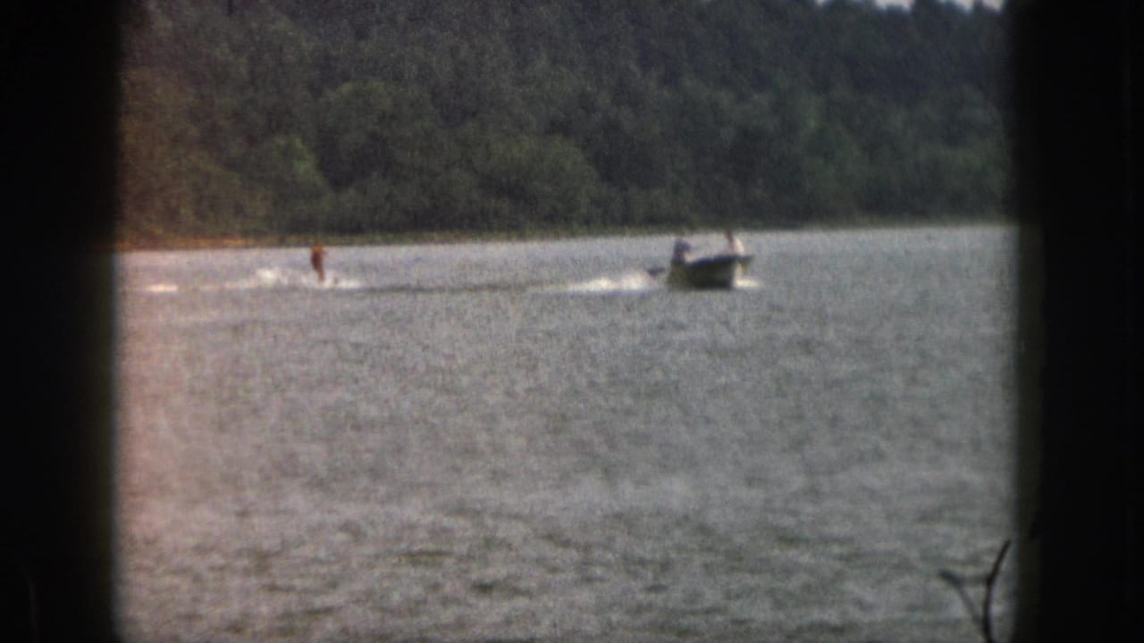 1961 Michigan 8mm frame showing a distant water skier being towed by a small motorboat on a tree-lined lake.