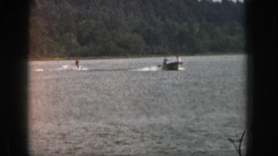 1961 Michigan 8mm frame showing a distant water skier being towed by a small motorboat on a tree-lined lake.