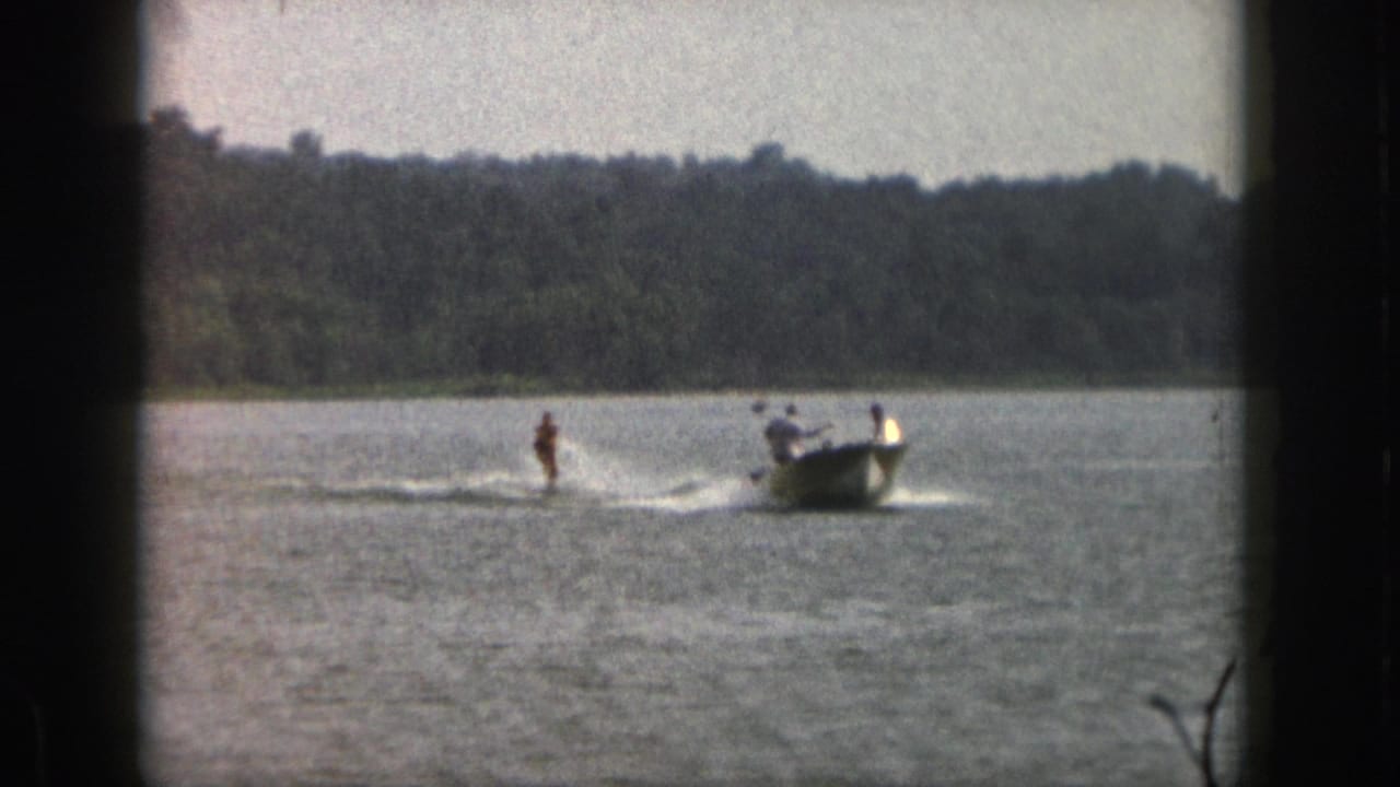 1961 Michigan lake scene with a water skier behind a small motorboat, framed by 8mm gate shadows and shoreline trees.