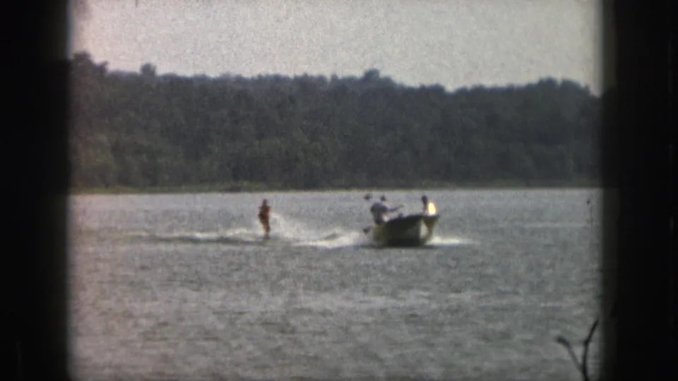 1961 Michigan lake scene with a water skier behind a small motorboat, framed by 8mm gate shadows and shoreline trees.