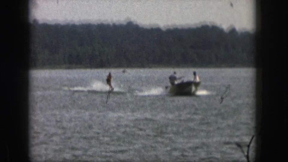 1961 Michigan 8mm frame of a water skier and small motorboat moving across a calm lake below a wooded shoreline.