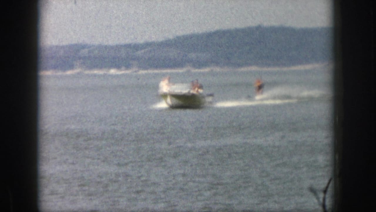 Soft 8mm frame from Michigan in 1961 with a boat towing a waterskier across a large lake under hazy light.