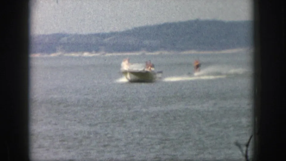 Soft 8mm frame from Michigan in 1961 with a boat towing a waterskier across a large lake under hazy light.