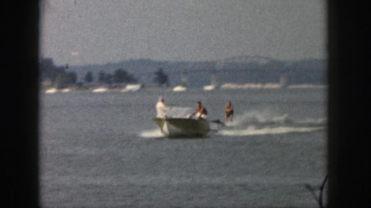 1961 Michigan home-movie frame showing a small motorboat towing two water skiers across calm lake water.