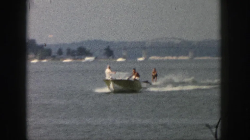 1961 Michigan home-movie frame showing a small motorboat towing two water skiers across calm lake water.