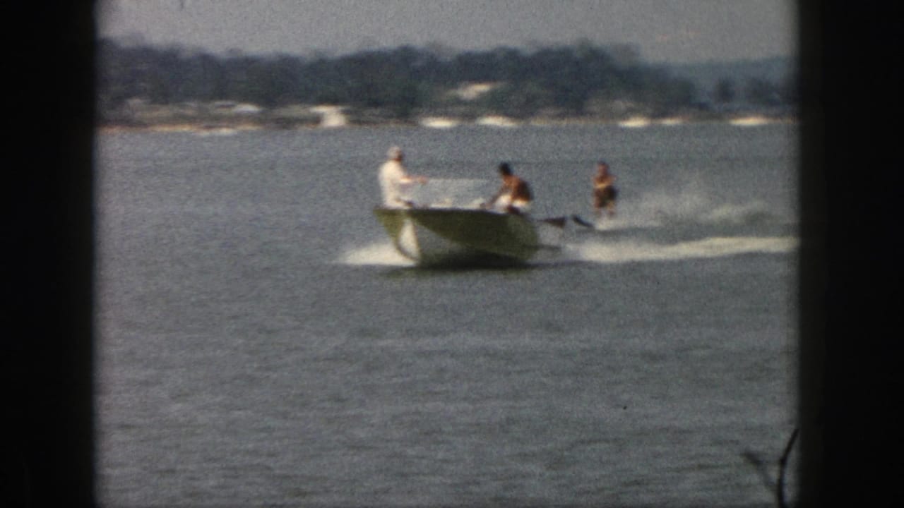 1961 Michigan lake scene with a green-and-white motorboat pulling two water skiers in soft 8mm color footage.