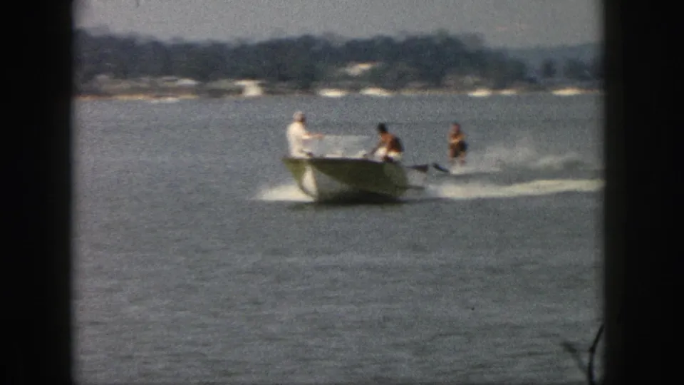 1961 Michigan lake scene with a green-and-white motorboat pulling two water skiers in soft 8mm color footage.