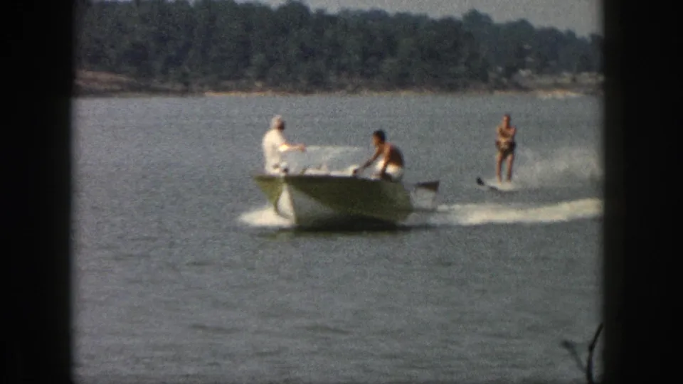 Closer 1961 Michigan home-movie view of a motorboat towing a standing water skier across the lake.