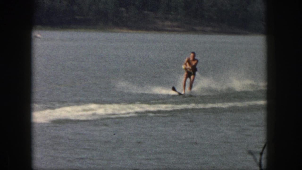 In 1961 Michigan home-movie footage, a shirtless man water skis across calm lake water behind an unseen tow boat.