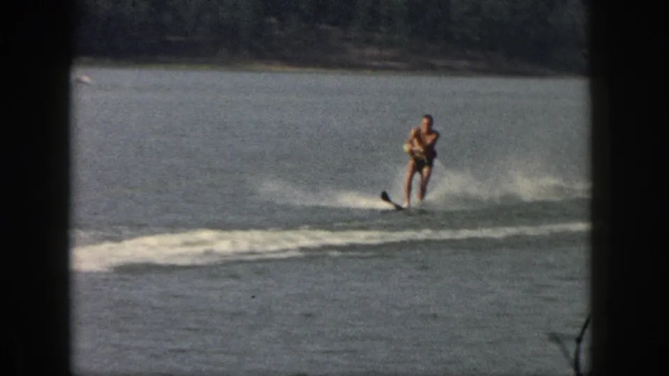 In 1961 Michigan home-movie footage, a shirtless man water skis across calm lake water behind an unseen tow boat.