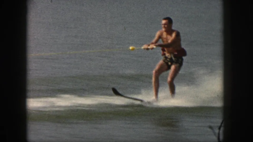 A closer 8mm frame shows the water skier in patterned trunks, knees bent and tow rope in hand, moving across the lake.