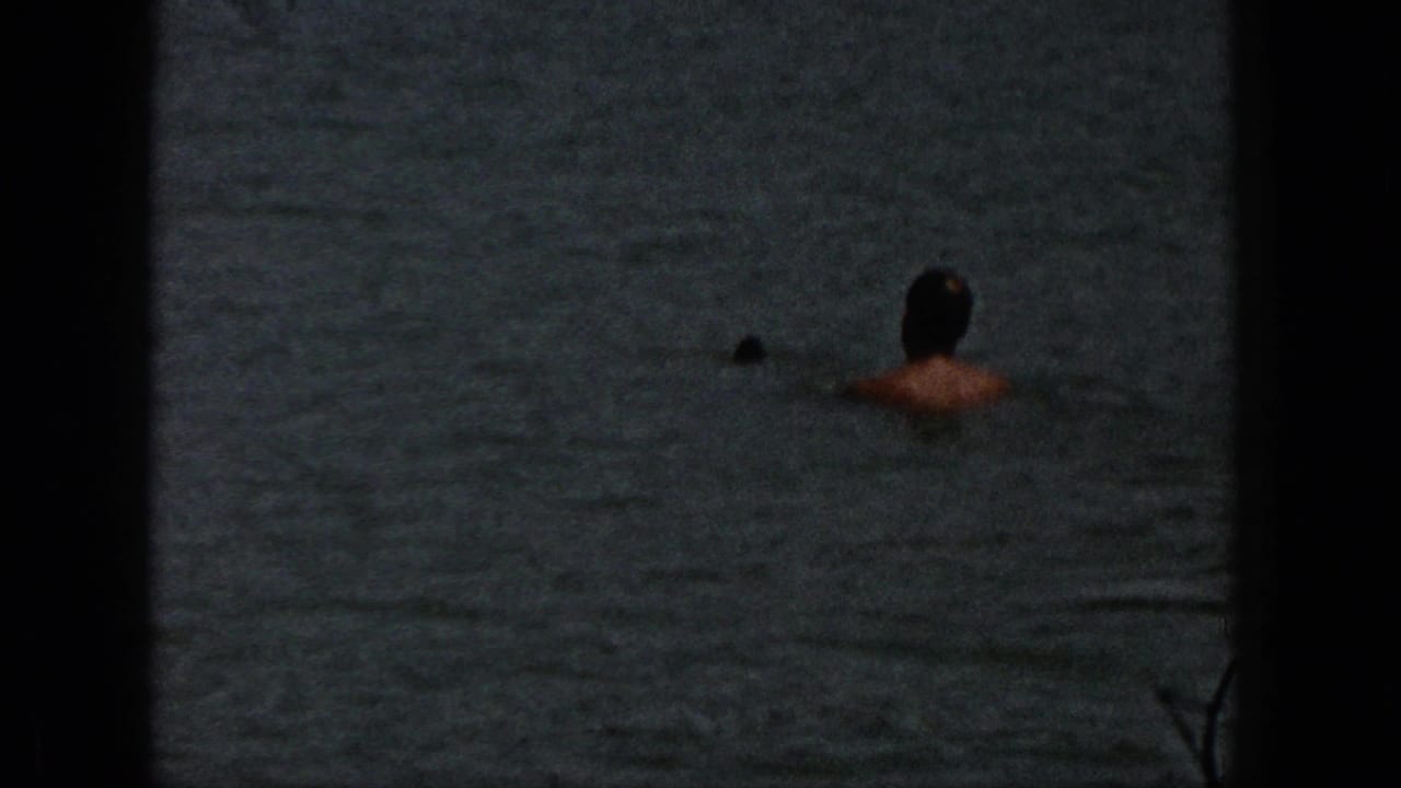 1961 Michigan home-movie frame showing a person in calm lake water near shore, partially obscured by dark 8mm frame edges.