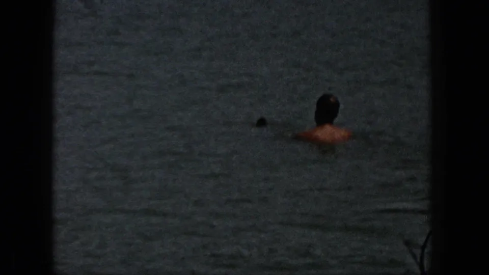 1961 Michigan home-movie frame showing a person in calm lake water near shore, partially obscured by dark 8mm frame edges.