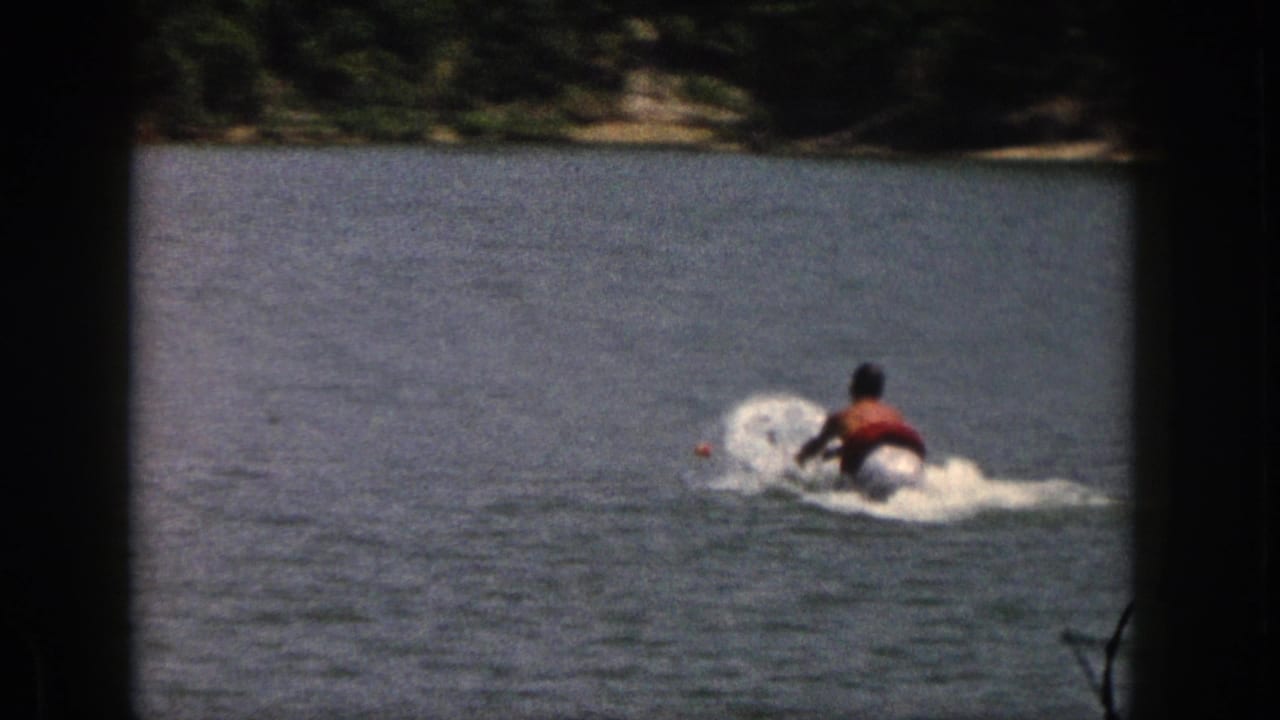 1961 Michigan home-movie frame showing a water skier moving across the lake with tow rope and spray, with trees along the far shore.