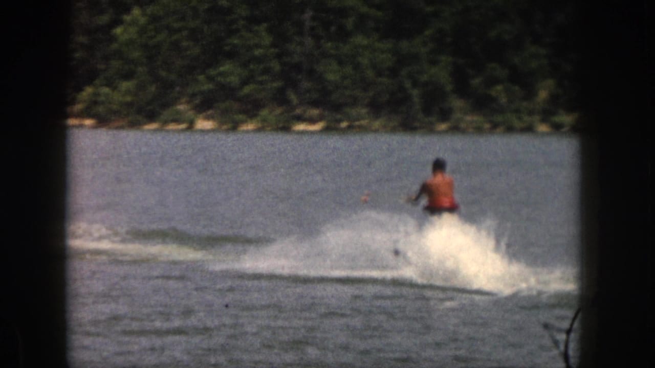 1961 Michigan frame showing a shirtless skier carving across calm lake water, with a broad white spray and tree-lined bank in the background.