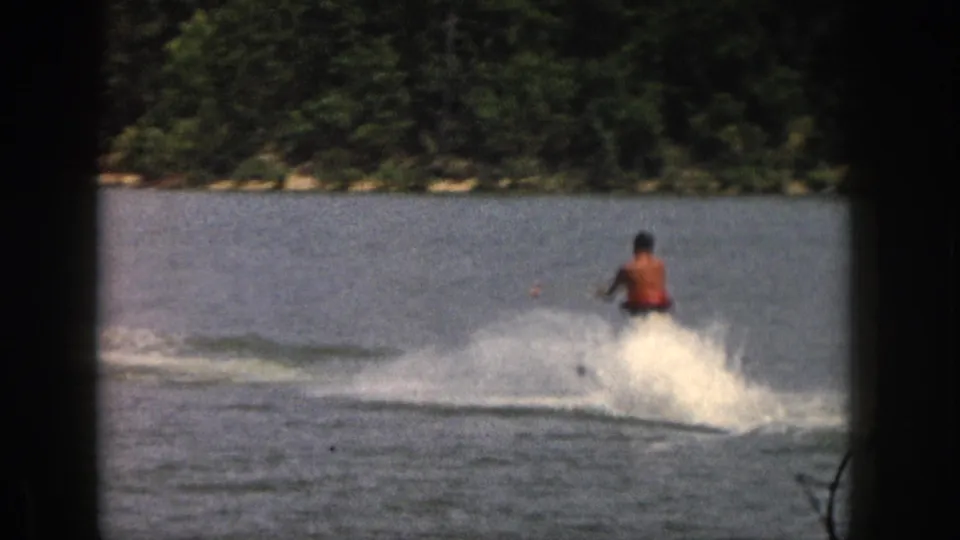 1961 Michigan frame showing a shirtless skier carving across calm lake water, with a broad white spray and tree-lined bank in the background.