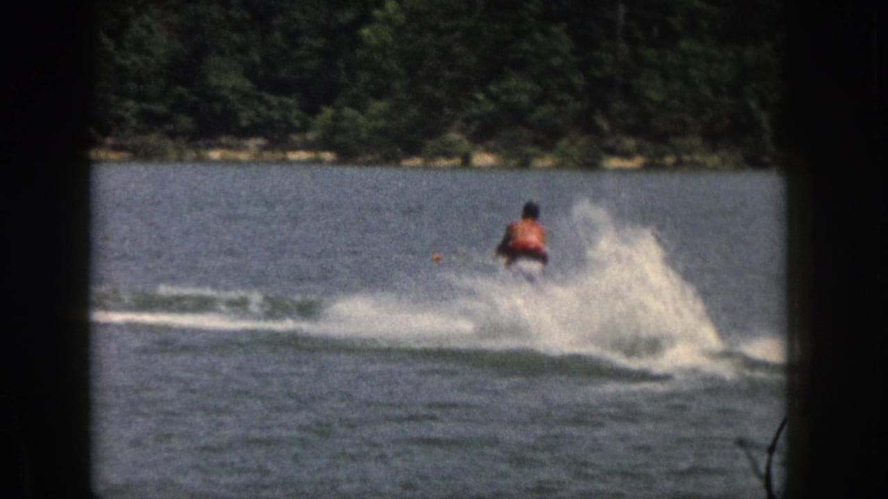 Water skier rides away across a lake, throwing white spray near a wooded shore.