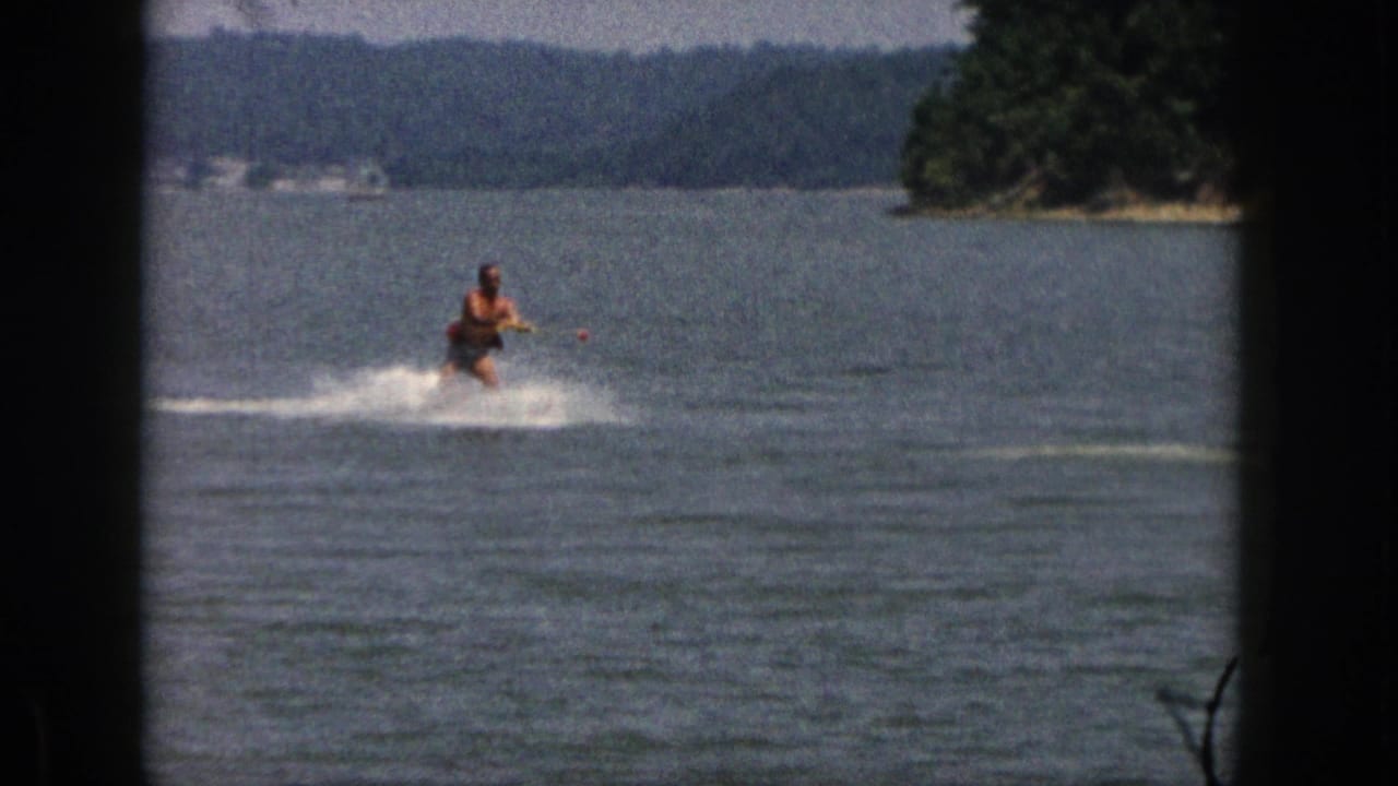 A distant adult skier rides toward camera across open lake water, with wooded Michigan shoreline and hills behind him.