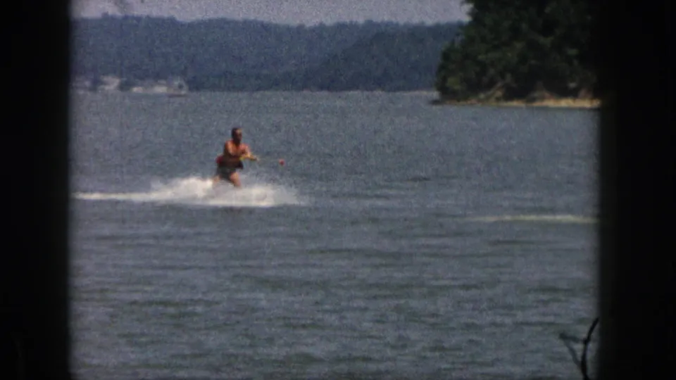 A distant adult skier rides toward camera across open lake water, with wooded Michigan shoreline and hills behind him.