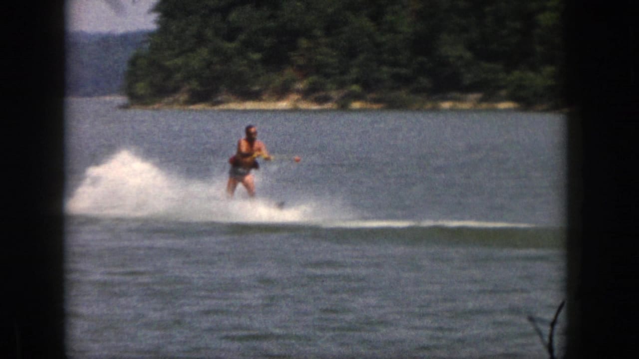 The adult skier glides nearer to the camera on a Michigan lake, holding the tow rope as a broad white spray rises behind him.