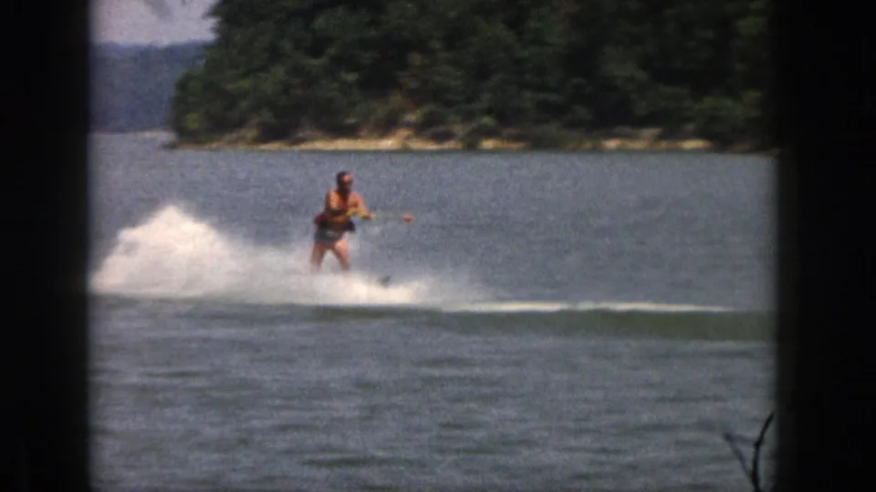 The adult skier glides nearer to the camera on a Michigan lake, holding the tow rope as a broad white spray rises behind him.