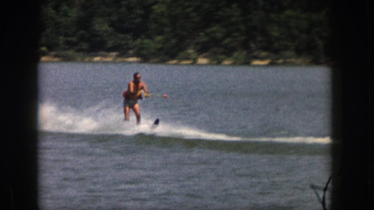 1961 Michigan home-movie frame of an adult man water skiing on a calm lake near a wooded shore.