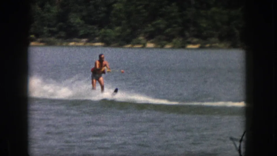 1961 Michigan home-movie frame of an adult man water skiing on a calm lake near a wooded shore.