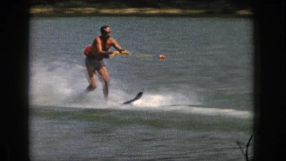 Closer 1961 Michigan frame of an adult water skier leaning forward while holding a yellow tow rope.