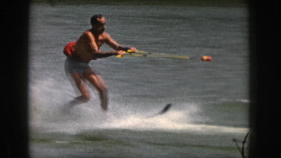 Adult water skier passes close in 1961 Michigan 8mm footage, holding a yellow tow rope over calm lake water.