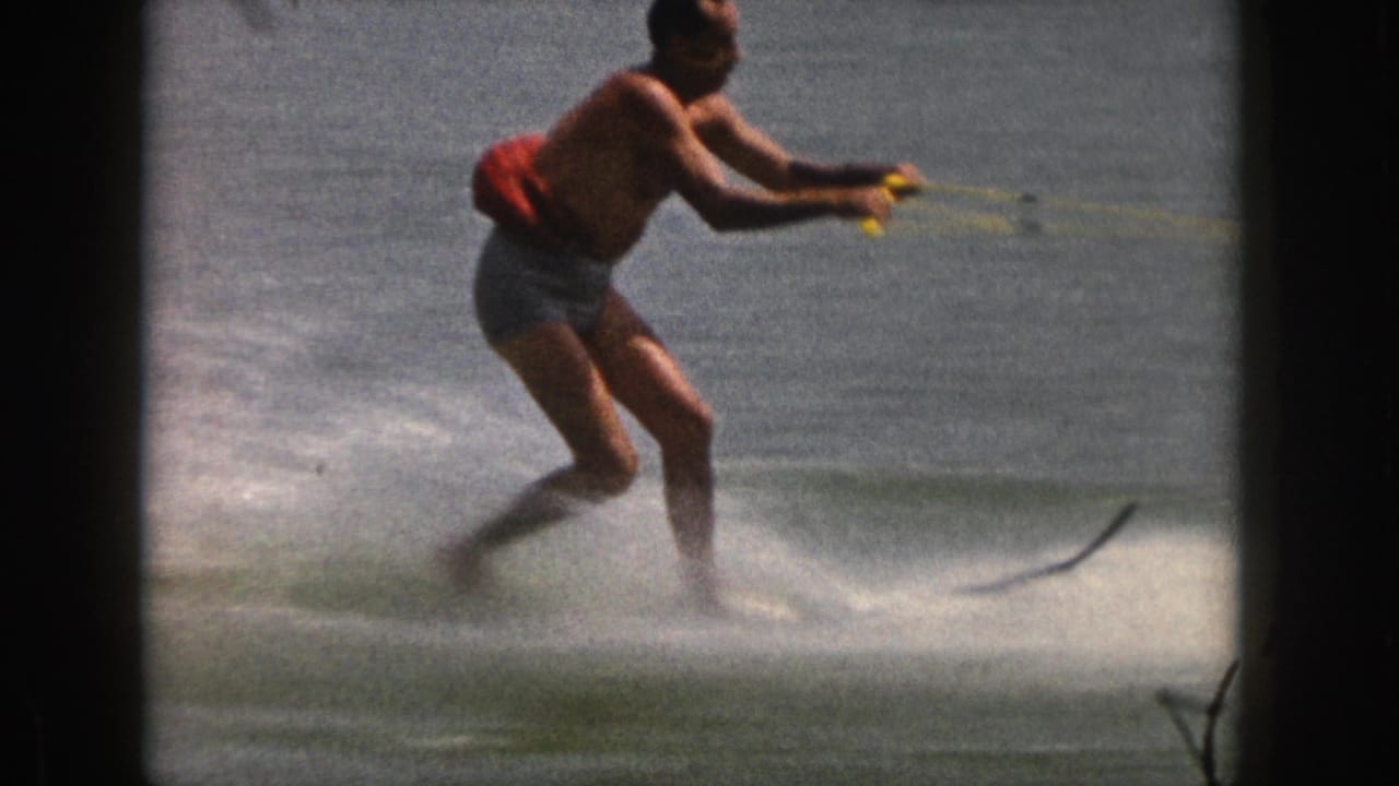 Man in a red life vest water skiing across a lake with spray beneath him.