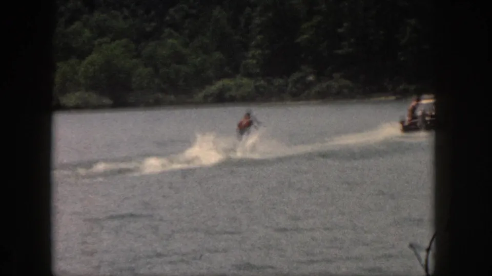 Wide 1961 Michigan 8mm view of a water skier being towed behind a small boat on a wooded lake.