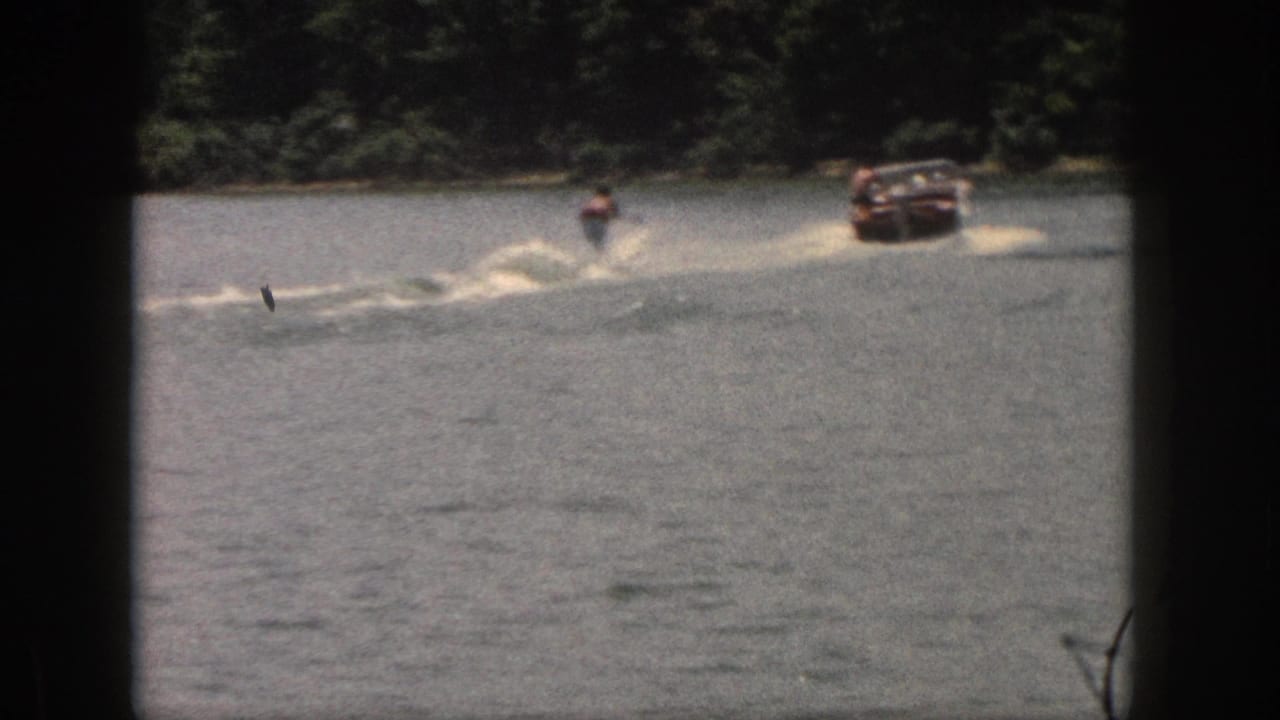 A motorboat pulls a distant skier across open water near a wooded shore.